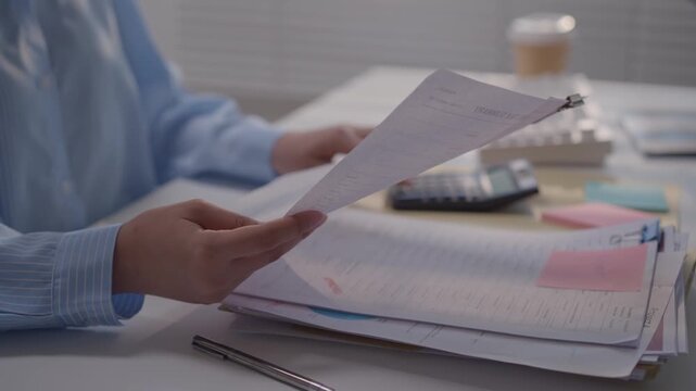 Person Reviewing Financial Document and Paper Invoice at Office Desk, Holding Paperwork Above Open Ledger with Calculator Nearby During Focused Routine Office Work
