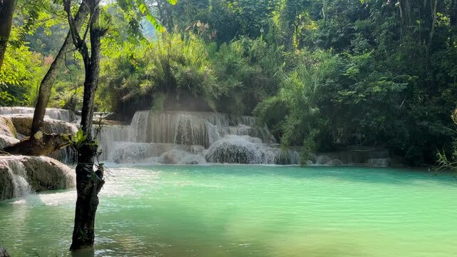 Turquoise pool framed by foreground trees at Kuang Si Waterfall near Luang Prabang, Laos, with tiered cascades and sunlit tropical forest.