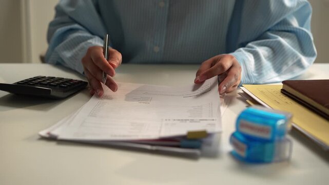 Person Reviewing Paperwork at Desk, Completing Document Forms and Finance Calculations with Pen, Writing Notes in Office Workspace Beside Calculator, Folder, Notebook, and Office Supplies