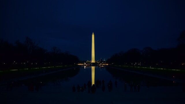 The lit Washington Monument and its long reflection align across the Reflecting Pool after dark, with trees silhouetted along both sides of the water. A winter view of Washington, DC, United States.