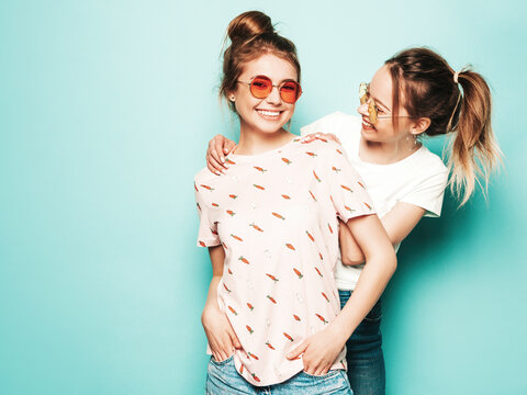 Two young beautiful blond smiling hipster in trendy summer hipster jeans clothes. Sexy carefree women posing near blue wall. Trendy and positive models having fun in sunglasses