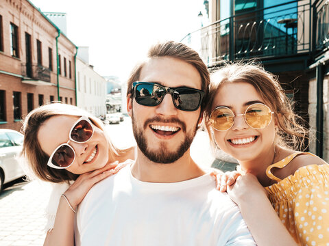 Group of young three stylish friends in the street.Man and two cute female dressed in casual summer clothes.Smiling models having fun in sunglasses.Women and guy making photo selfie on smartphone