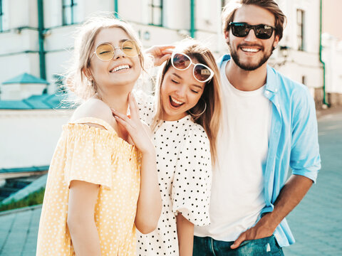 Group of young three stylish friends posing in the street. Fashion man and two cute dressed in casual summer clothes. Smiling models having fun near wall.Cheerful women and guy outdoors
