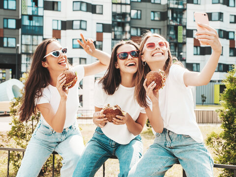 Three young beautiful smiling hipster female in trendy summer  clothes.Sexy carefree women posing in the street.Positive models taking selfie in sunglasses.Holding juicy burger and eating hamburger