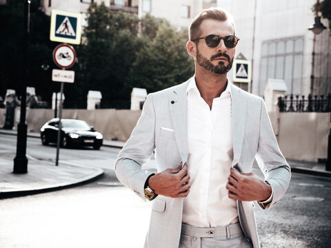 Portrait of handsome confident stylish hipster lambersexual model.Sexy modern man dressed in elegant white suit. Fashion male posing in the street background in Europe city at sunset. In sunglasses