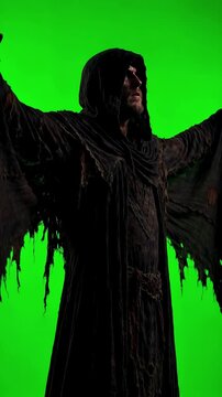 Slow dolly shot follows menacing hooded figure in tattered dark cloak raising arms slowly on uniform vivid green screen background with clean isolated edges