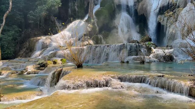 Main waterfall at Kuang Si near Luang Prabang, Laos, cascading over sunlit rock terraces into turquoise pools surrounded by tropical forest.