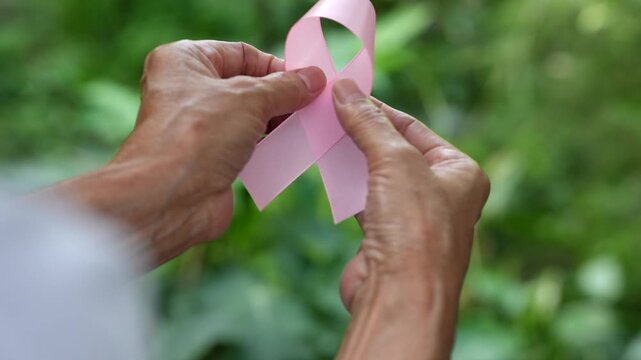 Close-up of older hands holding a pink ribbon outdoors against a soft green background, symbolizing breast cancer awareness, support, hope and survival.