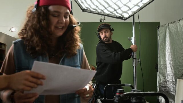 Caucasian young adult film crew adjusting studio lighting and reading script on production set. Ideal for video production marketing and backstage media projects