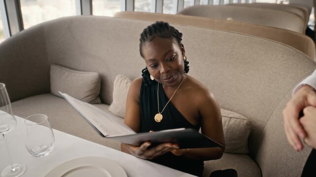 African American young adult woman receiving menu from waiter and ordering food in restaurant. Footage suitable for fine dining advertising, hospitality campaigns, and food service promotions