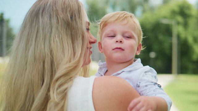 Mother gently kisses baby's forehead outdoors. Child looks curious and calm. Mother smiles warmly at her child. Scene captures tender, loving family moment. Great for parenting, child development