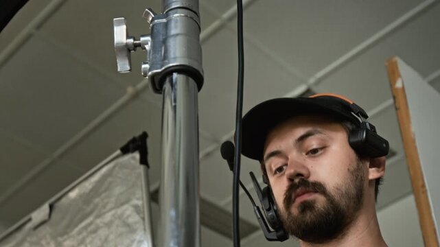 Caucasian young adult technician adjusting light stand and wrapping cables on film set. Suitable for backstage media production, filmmaking courses and cinematography videos