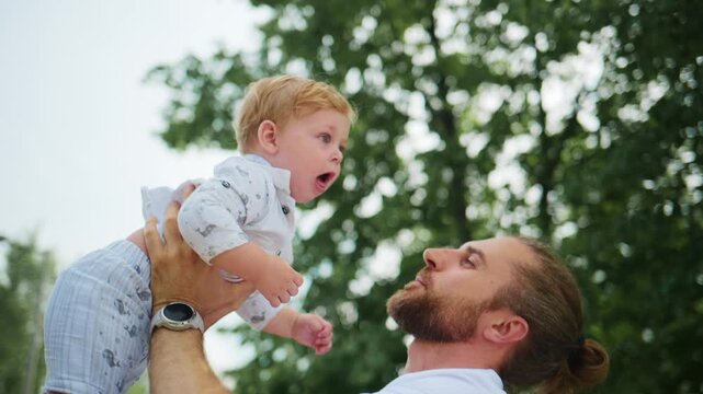Father joyfully lifts baby high in sunny park. Child giggles, arms spread wide. Dad smiles, eyes filled with love. Scene captures playful outdoor bonding moment. Perfect for parenting, family