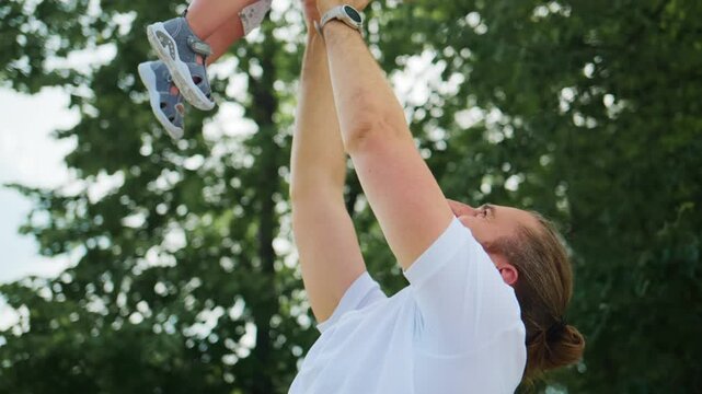 Man joyfully lifts toddler high into air outdoors. Father beams with delight as child laughs freely. Scene captures playful, carefree moment in sunny park. Perfect for parenting, family bonding