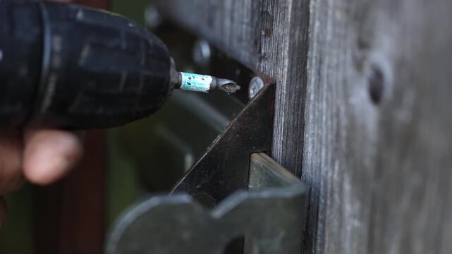 Close up view of worker driving screw into weathered timber fence using electric drill screwdriver, securing metal latch bracket with steady hands during garden gate repair work outdoors