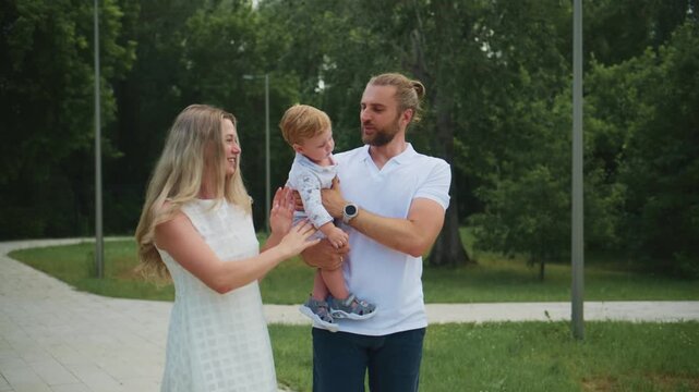 Young parents hold baby in park. Mother touches child's hand. Father looks at baby with affection. Family smiles warmly outdoors. Shot shows loving family moment in green space