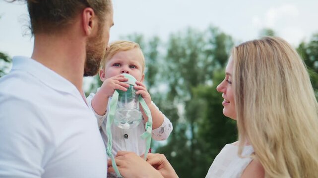 Young parents hold smiling baby outdoors. Baby grips green bottle with straw. Family laughs together under sunny sky. Perfect for parenting, family, or healthy lifestyle themes