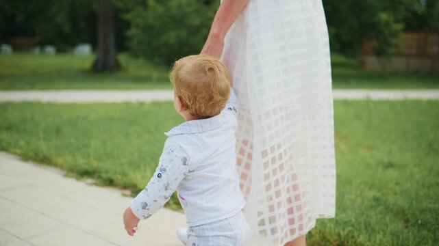 Little boy walks holding adult's hand. Mother walks beside child outdoors. Scene shows gentle care and family bond. Suitable for parenting, childhood, or wellness themes
