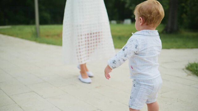 Young child walks with adult hand support. Child looks ahead with curious expression. Scene shows early walking milestone. Warm outdoor setting with greenery. Ideal for parenting, developmental
