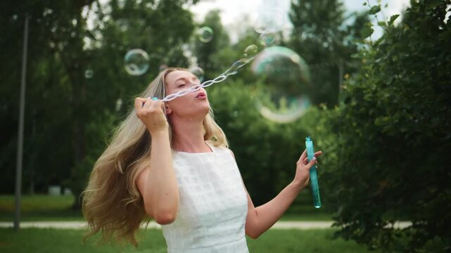 Young woman blows bubbles in sunny park. Her smile shows joy and relaxation. Bubbles float gently in green surroundings. Ideal for summer, happiness, or outdoor lifestyle themes