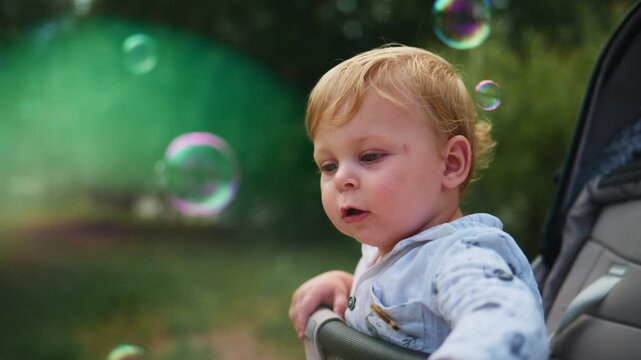 Young child sits in a stroller, eyes wide with wonder, chasing floating bubbles. Soft sunlight highlights golden hair as he reaches out, curious and delighted. Greenery surrounds him