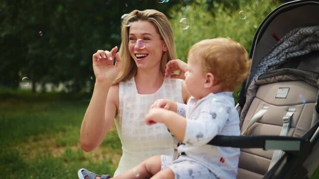 Mother blows bubbles while baby in stroller watches with wonder. Warm, joyful moment in sunny park. Perfect for family, parenting, or outdoor lifestyle themes