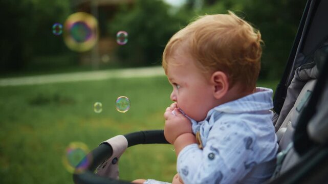 Baby in stroller watches floating bubbles with curiosity. Child's hands near mouth, eyes focused on bubbles. Soft outdoor lighting, green grass background. Ideal for parenting, childhood, outdoor play