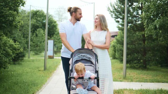 Happy family walks together in a park. The parents smile while pushing a stroller with a toddler. They stroll along a grassy path surrounded by green trees. Ideal for scenes of family bonding
