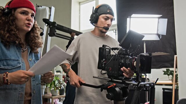 Caucasian young adult woman and man operating professional cinema camera while discussing script on studio set. Ideal for film production, creative media agency, and cinematography advertising