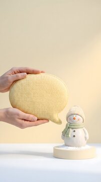 Hands holding a yellow knitted speech bubble sign next to a cute snowman figurine wearing a matching winter hat and scarf against a neutral background