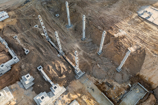 An aerial view shows concrete piles and foundation footings across a building site, with soil excavation and construction materials visible around the lot