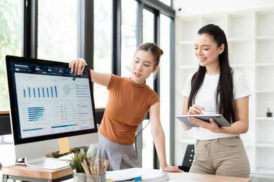 Two Asian Businesswomen Discussing Financial Data on Computer Monitor and Tablet