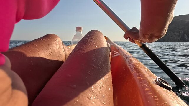 Paddle athlete navigating orange canoe near dramatic rock cliff. First-person perspective capturing repetitive motion on shimmering ocean surface.
