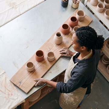 High angle view of a female potter wearing an apron and examining handmade ceramic vases and cups on a wooden board in a bright pottery studio