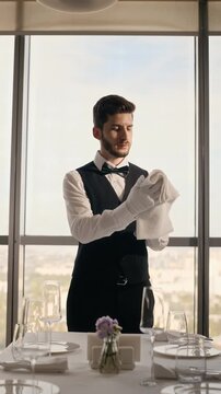 Young adult Caucasian waiter polishing wine glass with napkin and setting table in luxury restaurant. Clip ideal for hospitality training and catering advertising, vertical