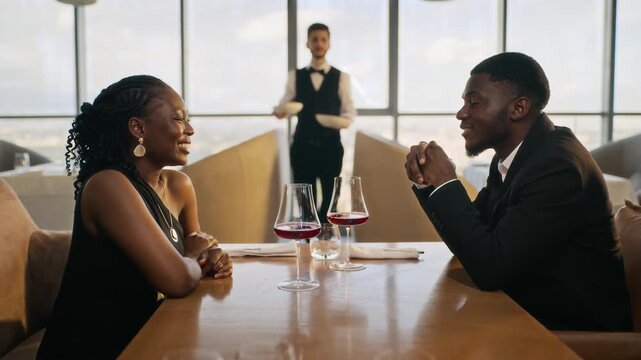 Young adult African American couple talking during romantic date while waiter serving gourmet dishes at restaurant. Suitable for fine dining advertising, dating service promos and luxury marketing