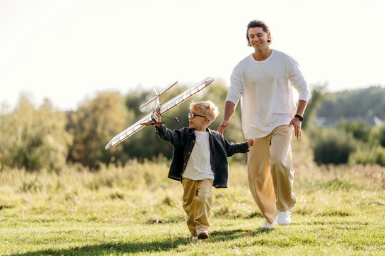 Adult and child running together through sunlit grassy meadow with toy airplane, joyful outdoor family scene in a natural rural landscape