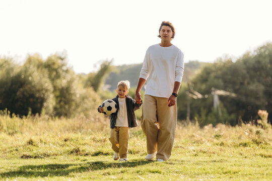 Adult man and young child walking across a sunlit grassy field while holding a soccer ball in a peaceful outdoor landscape