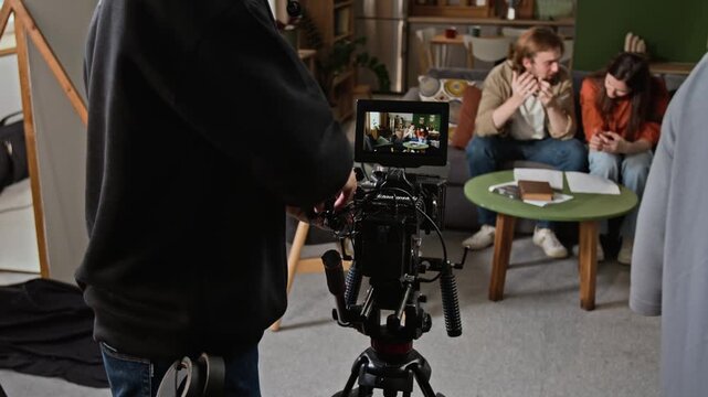 Young adult cameraman operating cinema camera rig from rear view while filming actors. Clip suits video production marketing and film industry projects