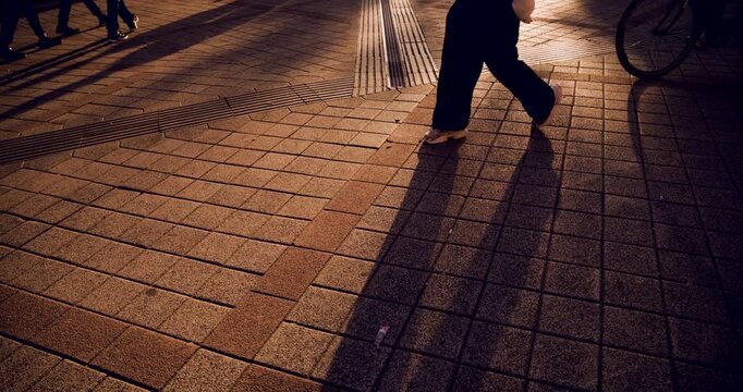 Cyclists and pedestrians passing from right to left across a brick paved city sidewalk, with long shadows and warm golden evening light and a tactile paving strip on the ground. 4K.
