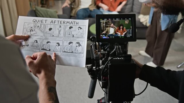 Young adult filmmaker holding storyboard and discussing scene near camera monitor while actors rehearse on set. Footage ideal for video production, media education, and cinema industry promos