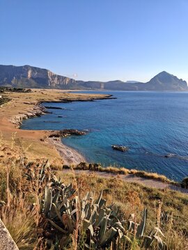 Veduta panoramica di una costa caratterizzata da vegetazione secca e rocce affacciate sul mare, con montagne visibili all'orizzonte.