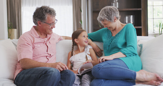 Happy caucasian grandparents and granddaughter sitting on sofa, reading book together