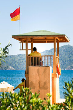 A lifeguard on duty sitting in a wooden observation tower at a tropical beach resort, watching over swimmers. Red and yellow safety flag waving above the tower