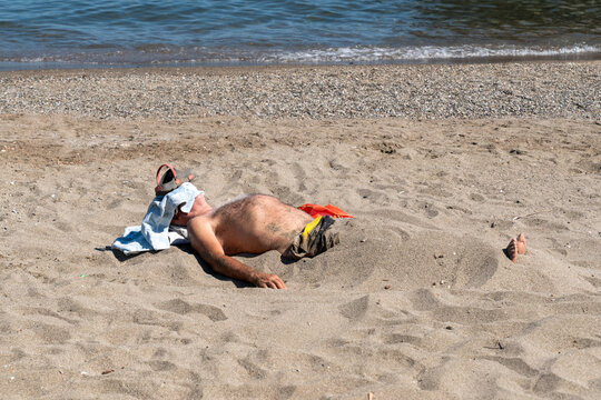 A senior man buried under warm sand relaxing and sunbathing on a sandy beach by the sea shore. 