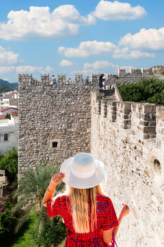 Back view of a blonde female tourist wearing a red dress and white sun hat, exploring the historic stone walls and towers of Marmaris Castle in Turkey. 