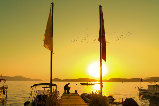 Golden sunset over Sogut village harbor with man sitting on wooden pier, Turkish flag and flying birds, Marmaris, Turkey
