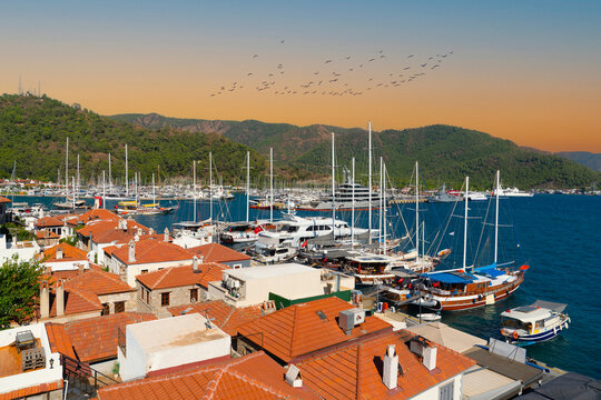 High angle view of Marmaris marina with yachts, red tiled roofs and birds flying under warm sky, Mugla, Turkey