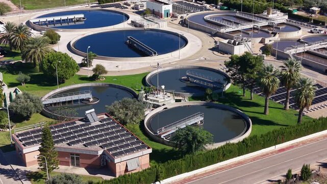 Wastewater treatment plant with circular clarifier tanks, rotating scraper bridges and rooftop solar panels surrounded by palm trees and green lawns
