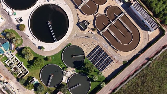 Aerial view of a wastewater treatment plant with brown aerated reactor basins, circular clarifiers and a ground-mounted solar array in Spain